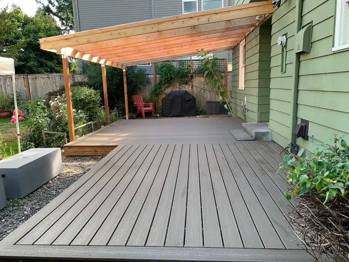 Gray composite deck under a cedar pergola beside a green house