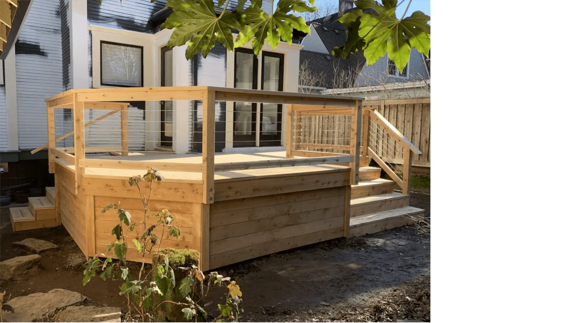 Elevated cedar deck with horizontal stainless-steel cable railing, attached to a white century home, shown on a sunny day.